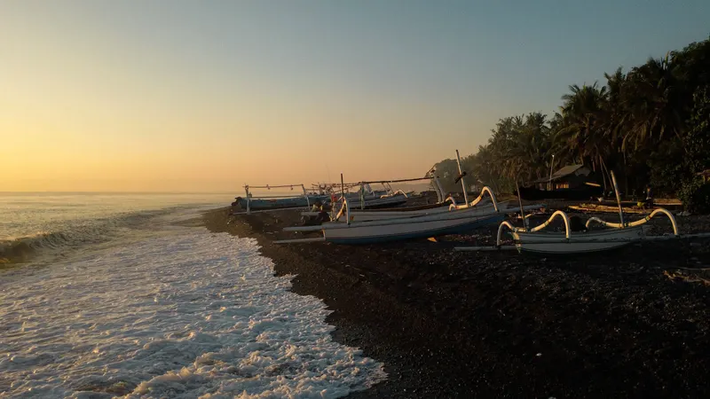 Outrigger fishing boats drawn up on Tianyar's pebble shore with the sun low over the water, palms lining the coast behind them