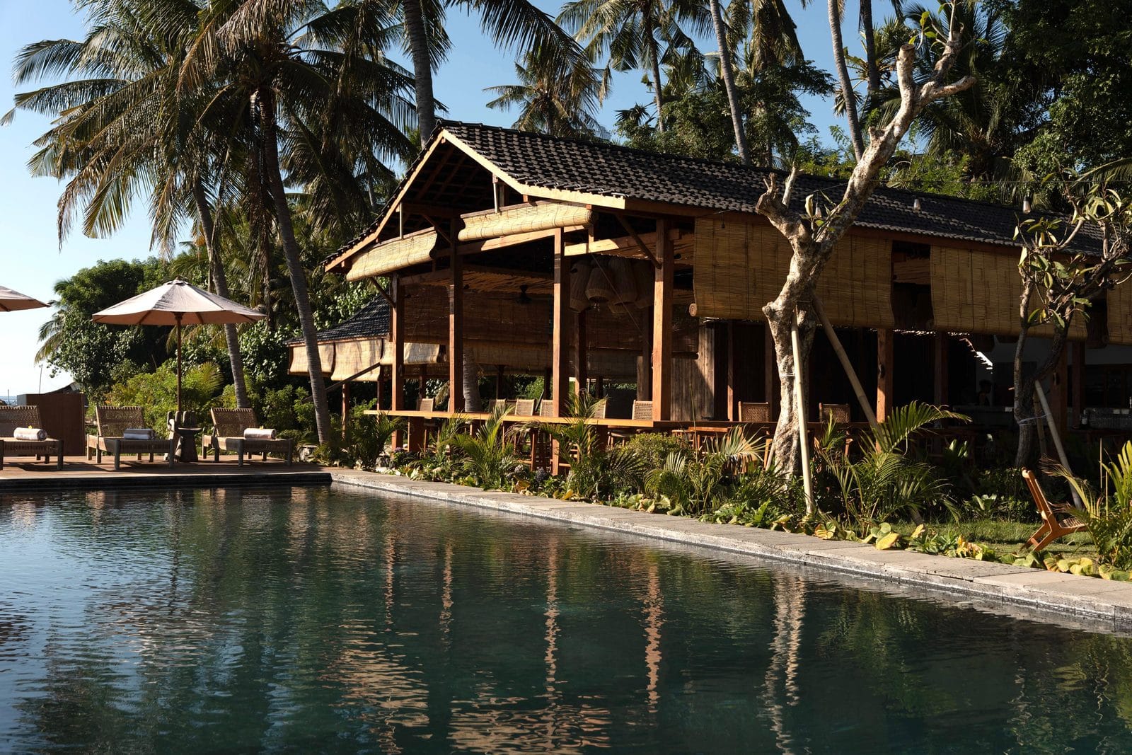 The open kitchen pavilion at Saga beside the pool, bamboo blinds rolled down, rattan chairs under the roof and coconut palms behind it in the morning sun