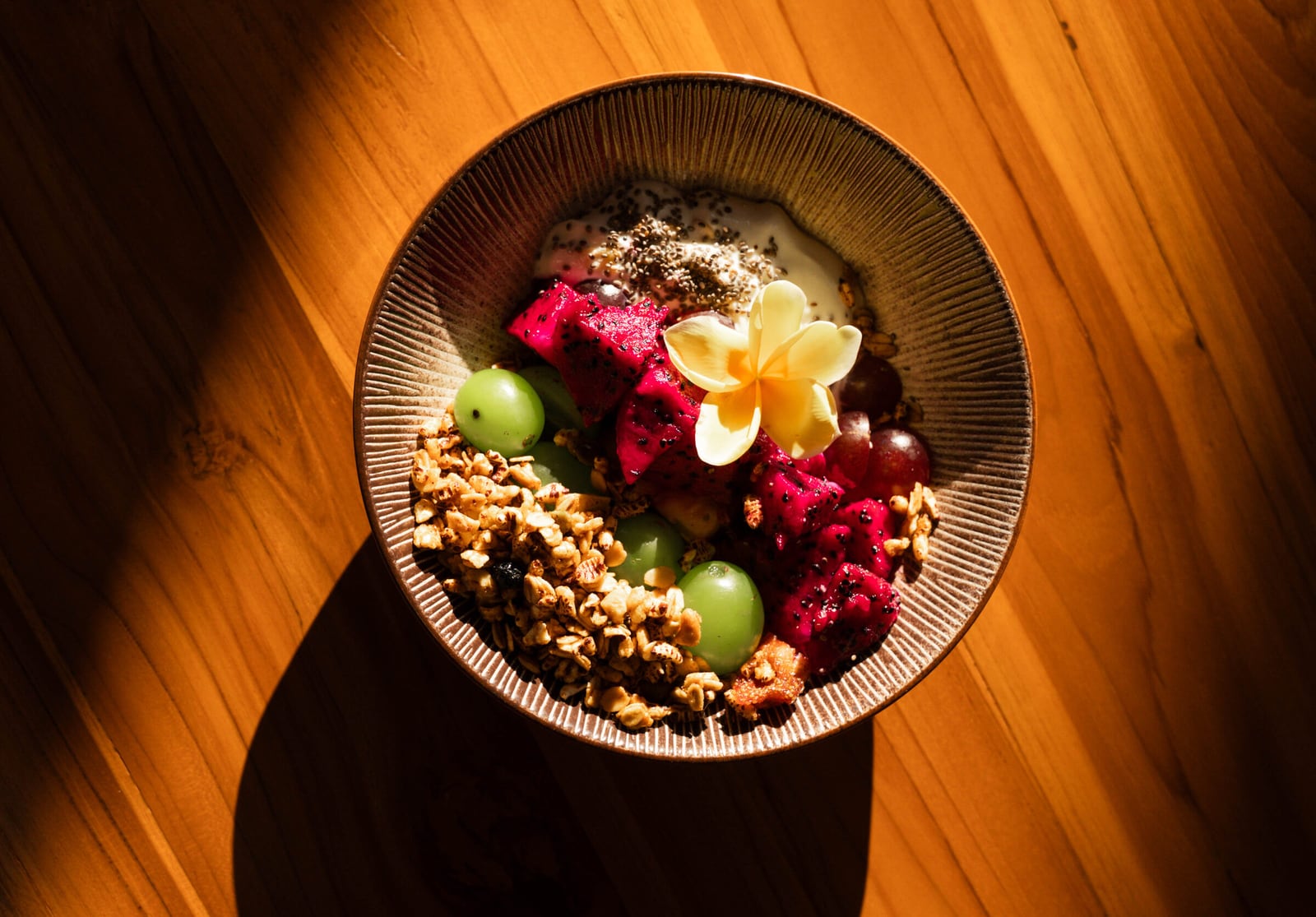 A breakfast bowl with dragon fruit, grapes and granola on a teak table in morning light