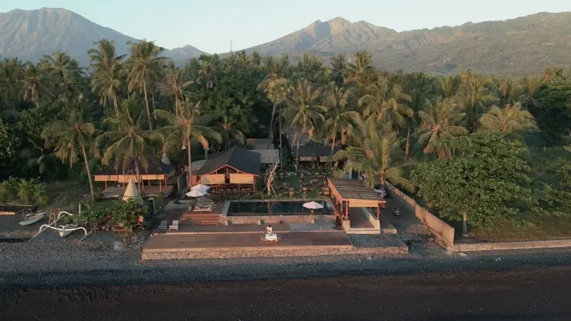 VELA Tianyar from the sea at low sun: the pebble shore, the pool and Saga at the water, the lodges behind under the palms, Mount Agung beyond