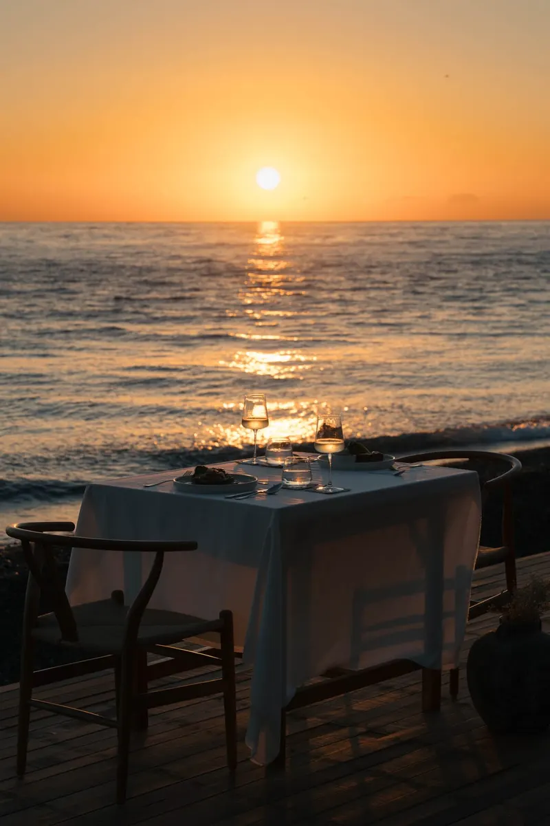A table laid for two on a timber deck at the shore, two glasses on a white cloth, the sun low over the sea